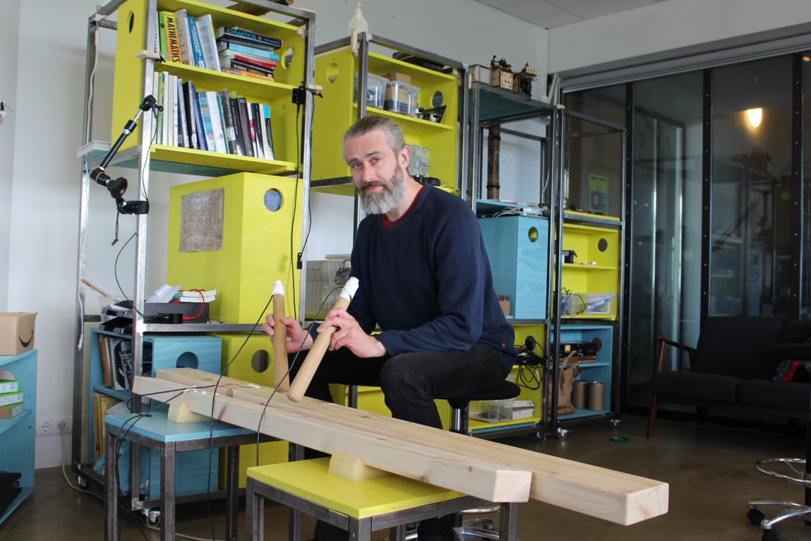 Man sitting in front of a wooden percussion instrument, holding large wooden sticks. Yellow and blue shelving system in the background.