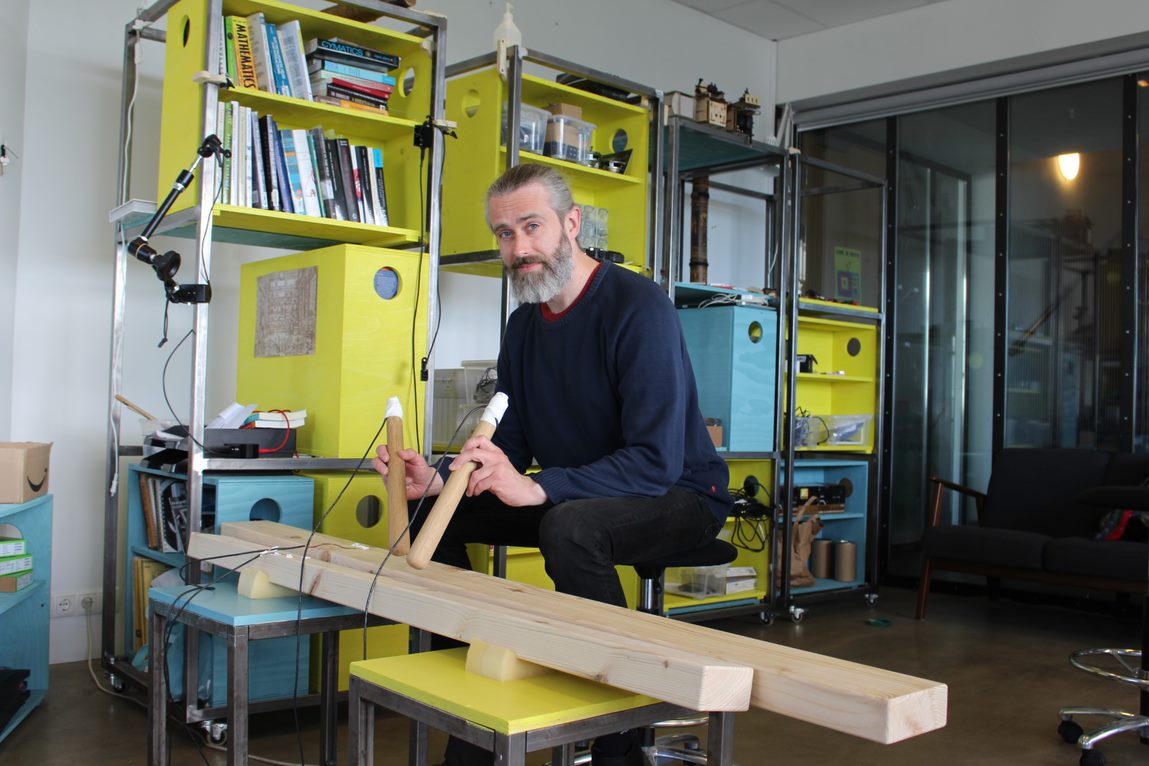 A man playing txalaparta, in the background a yellow shelving system.