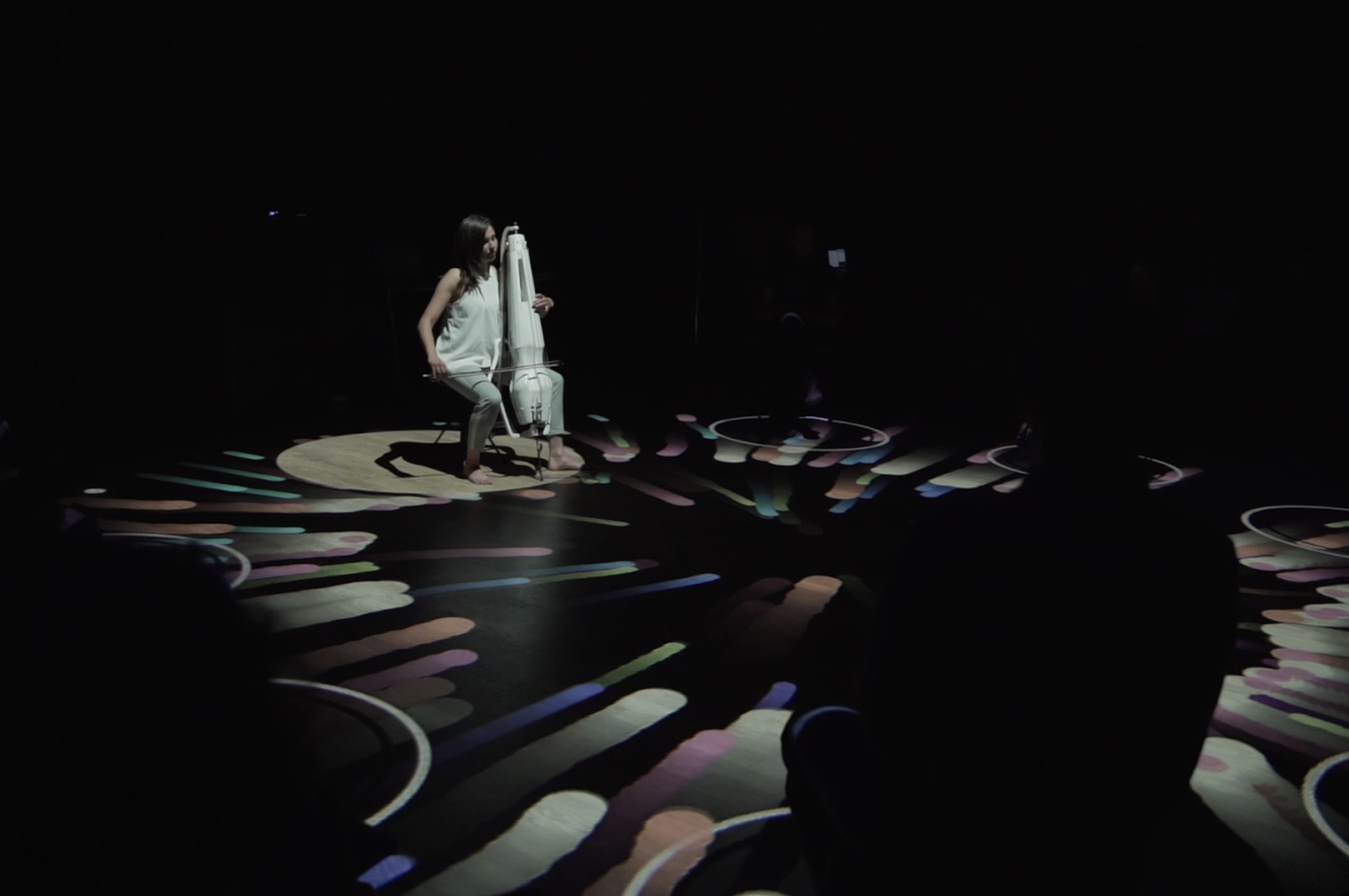 A young woman playing the knurl like a chello, in a middle of a room with colorful patterns on the floor and sitting audience in a circle around her.