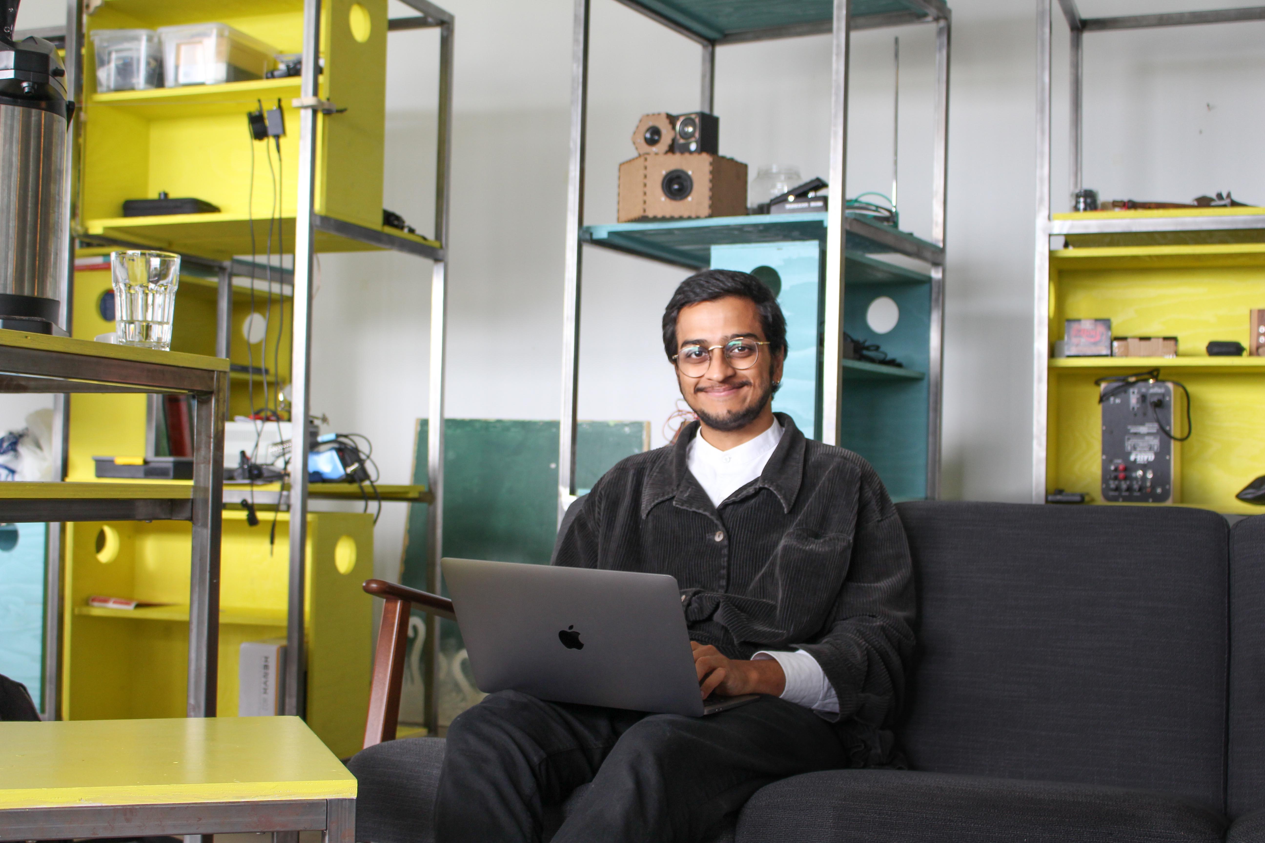 Young man sitting in a gray couch with a laptop, in front of a yellow shelving system.