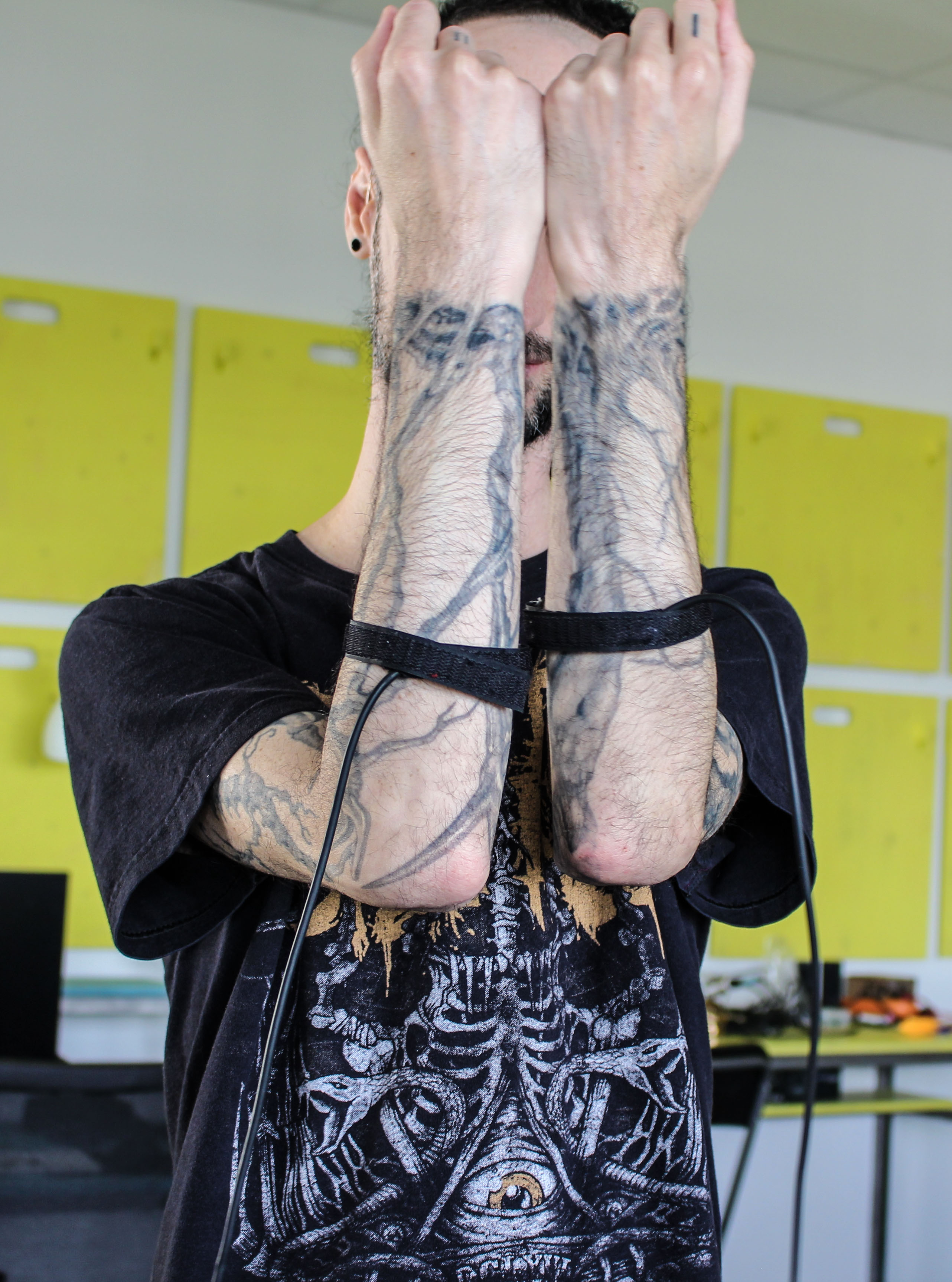 A person holding tattoo covered arms in front of their face, sensors are attached to their arms.