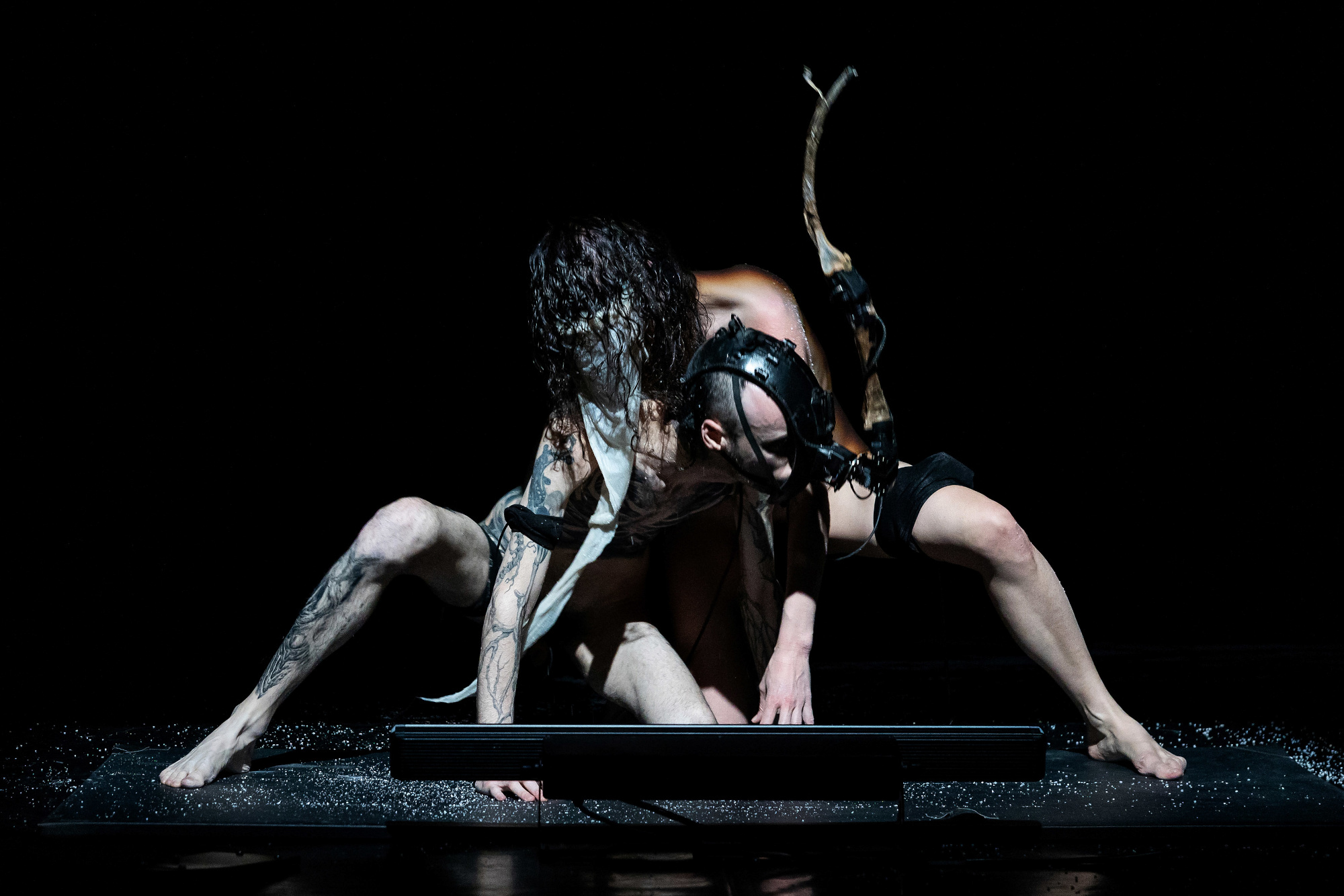Two performers on a black stage, dramatic dancing close to each other on the floor. Both have covered faces, one with a prosthetic and the other one with cloth.