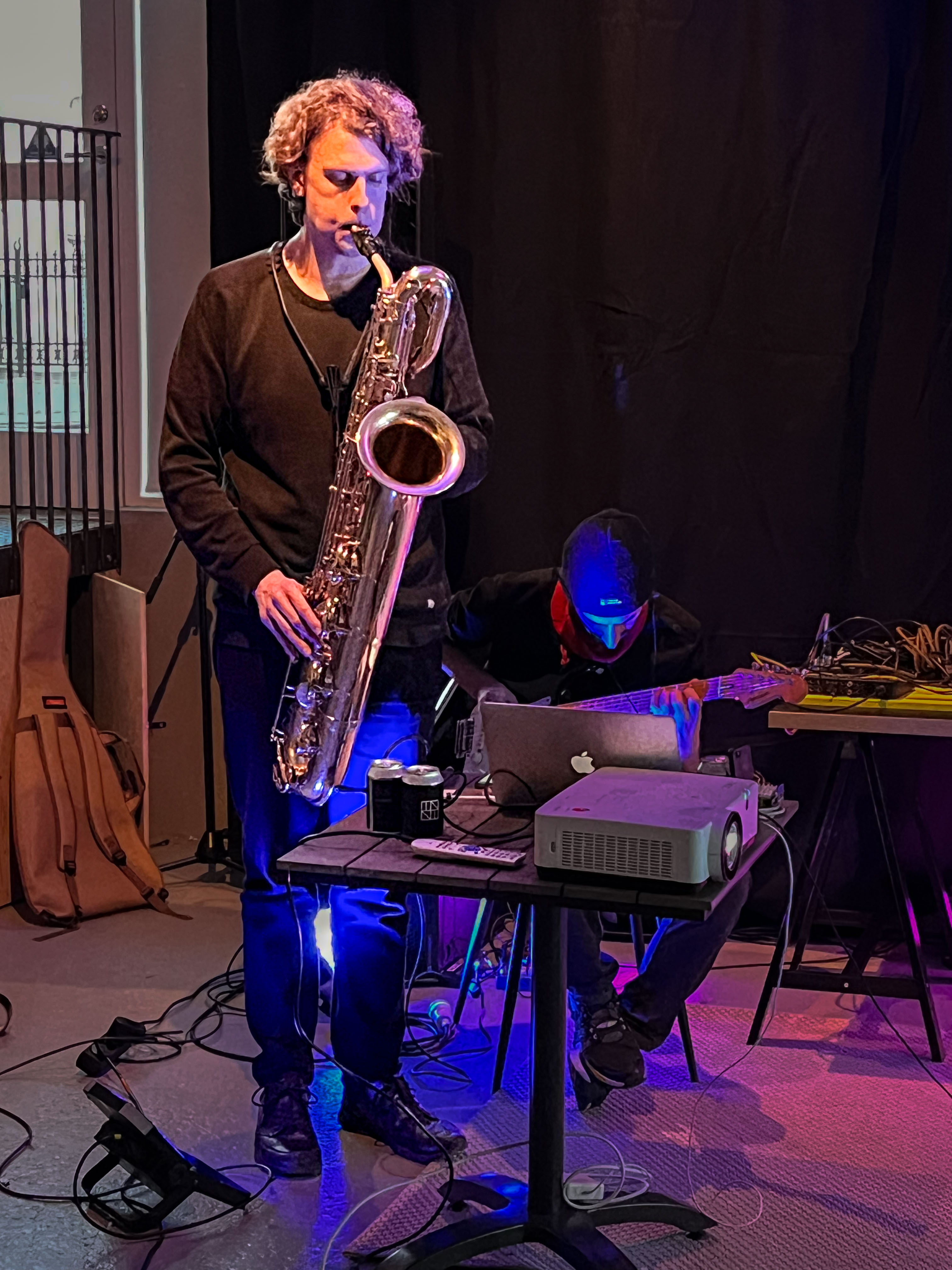 Two men playing music together, one standing with a saxaphone and a headband, the other one sitting in front of a laptop with an electric guitar.