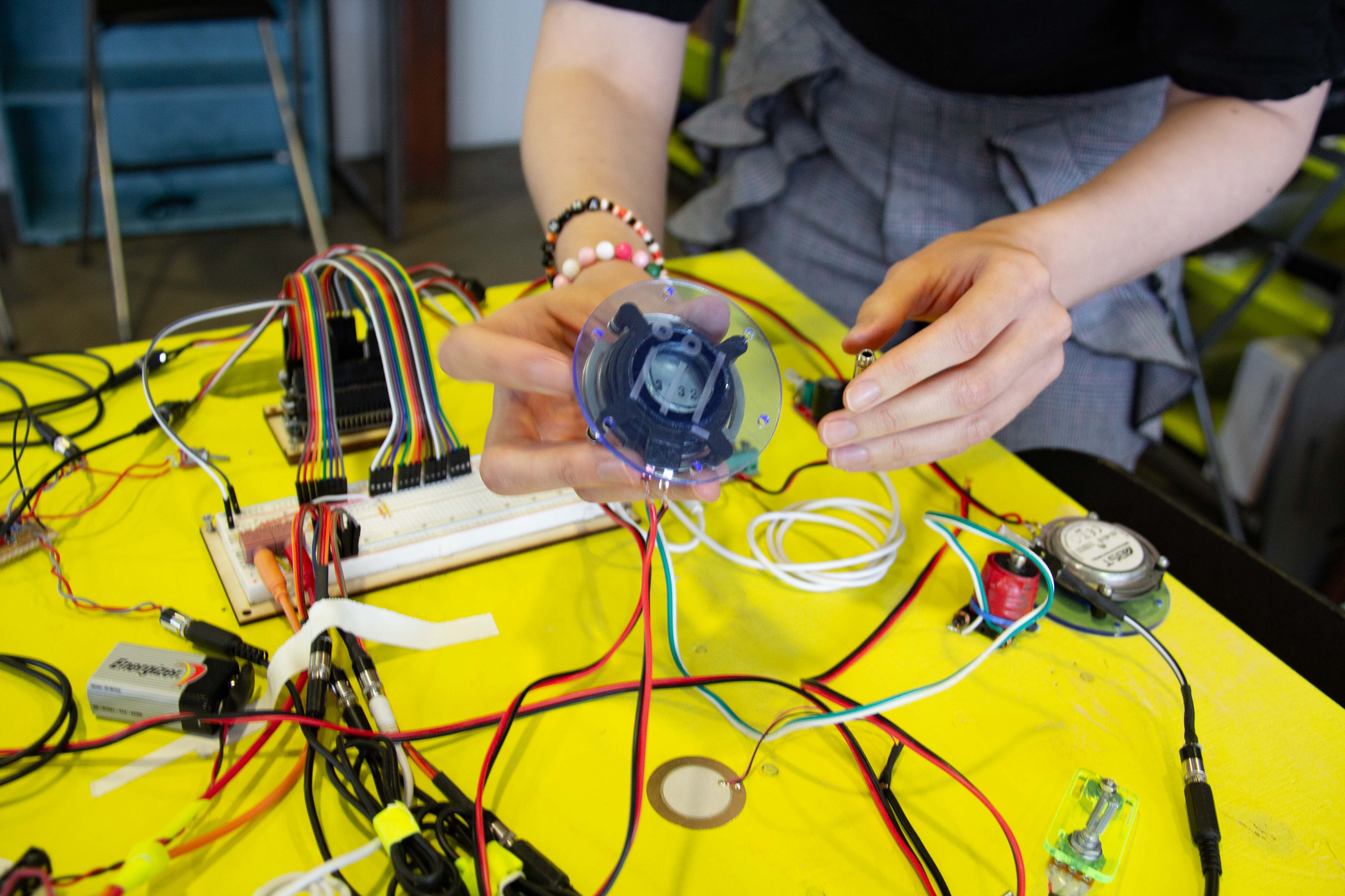 Various electronics on a yellow table, hands showing a speaker