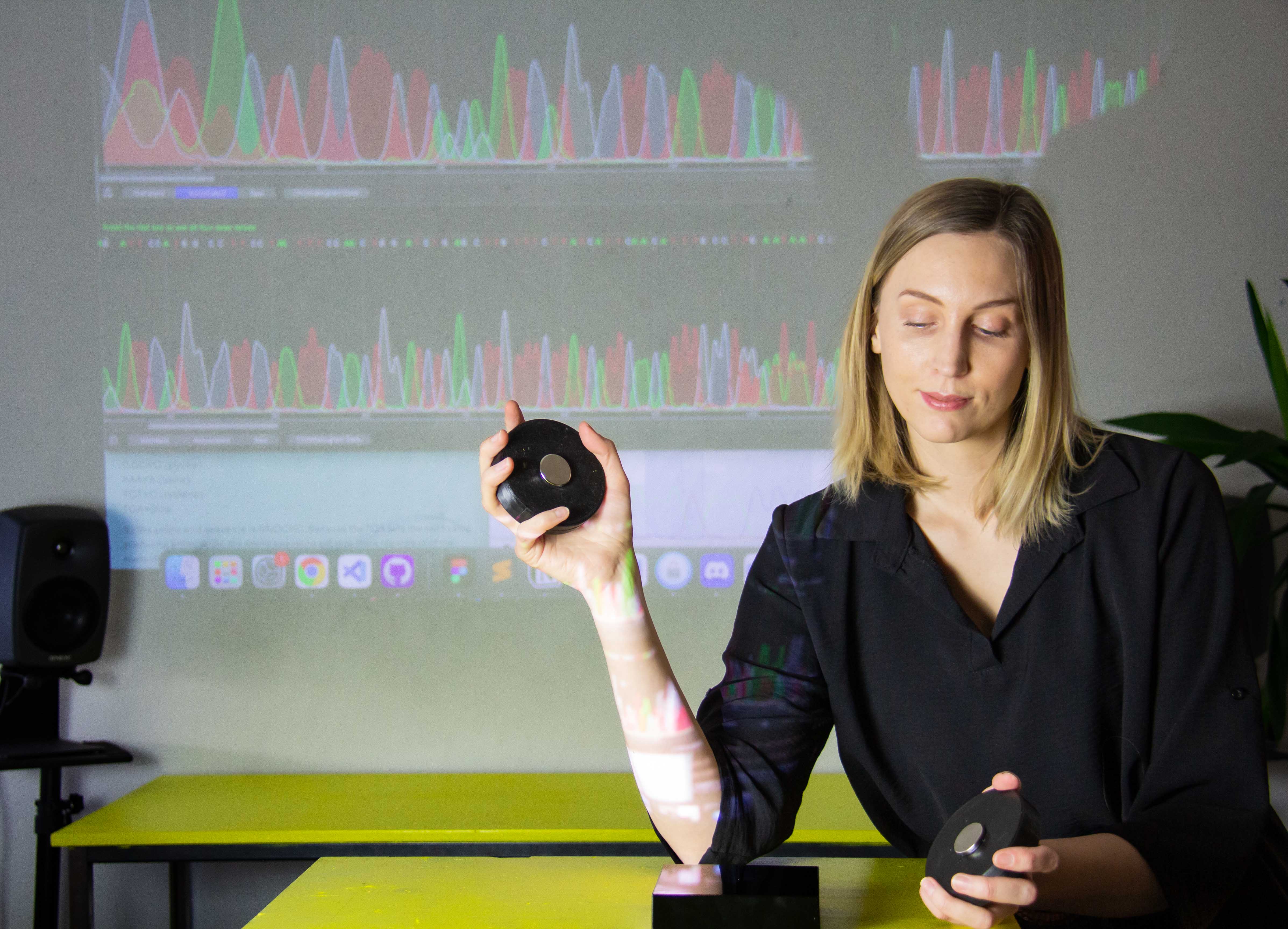 A smiling young woman holding a magnetic controller. In the background there is a screen with visualisation of data.