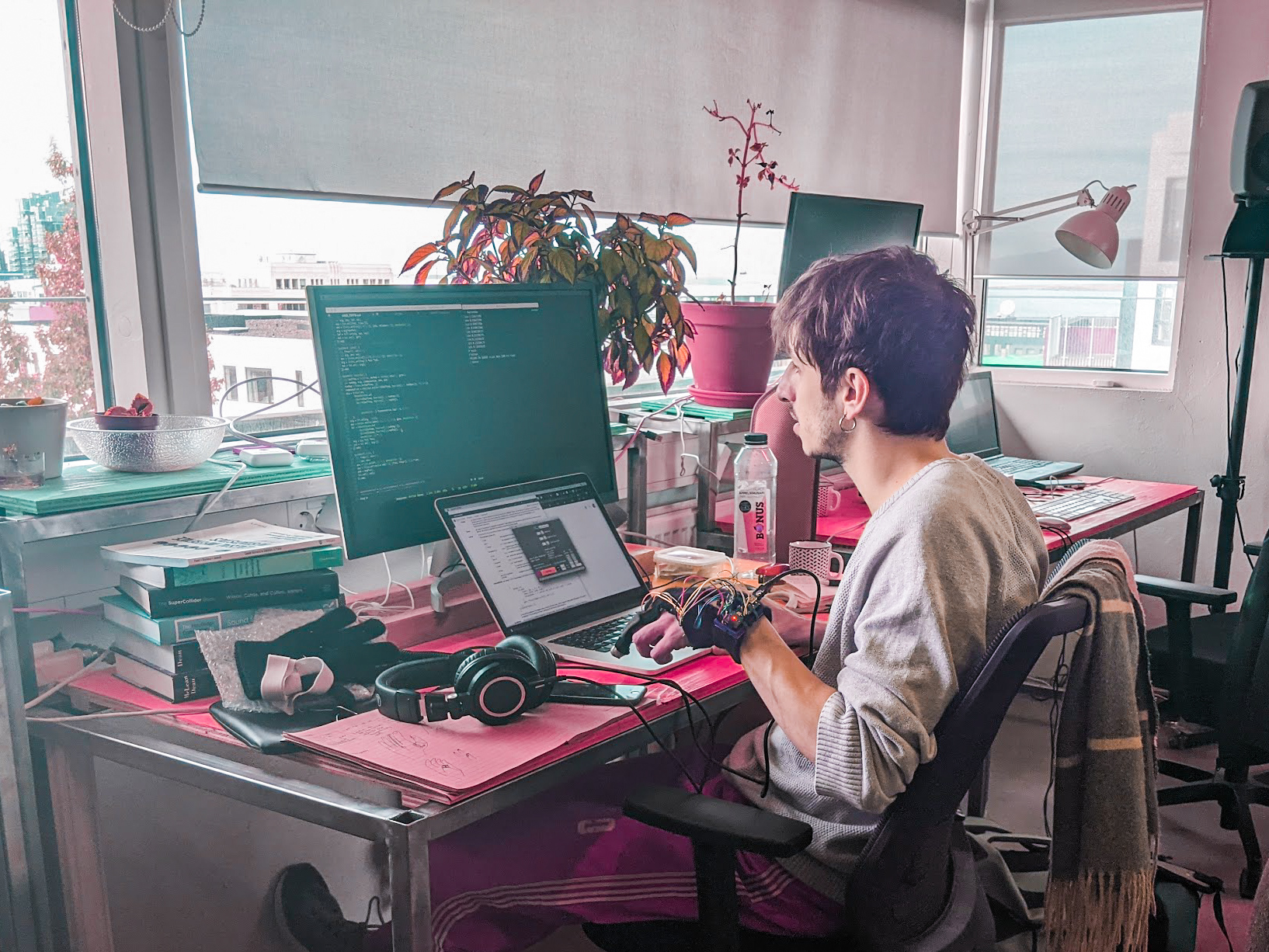 A young person sitting in front of a laptop and a computer monitor by a pink desk filled with books and technical elements, on their hand is an electronic glove with loads of chords plugged in
