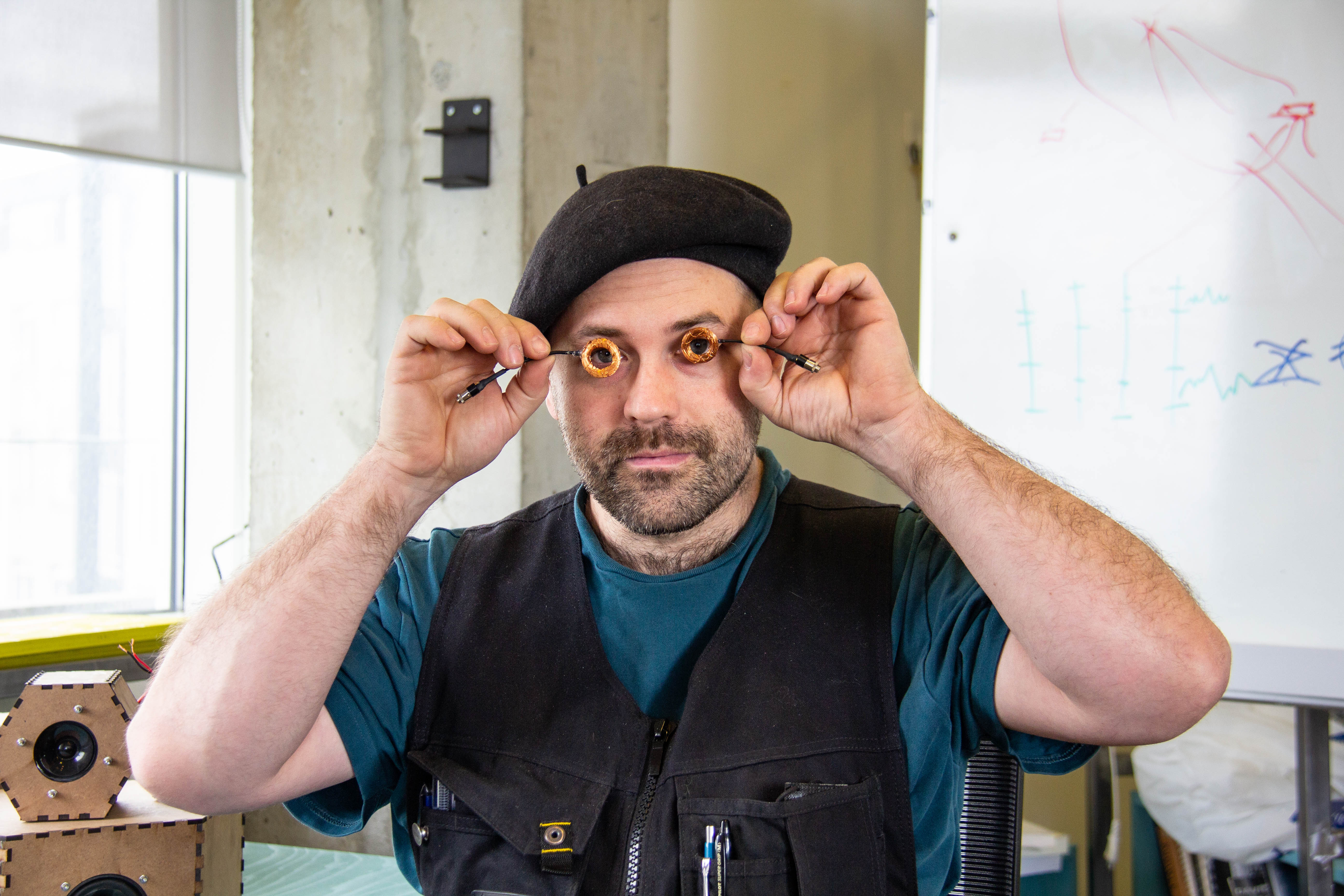 A man sitting at a blue desk, peeking through a couple of tiny bronze circles he's holding.