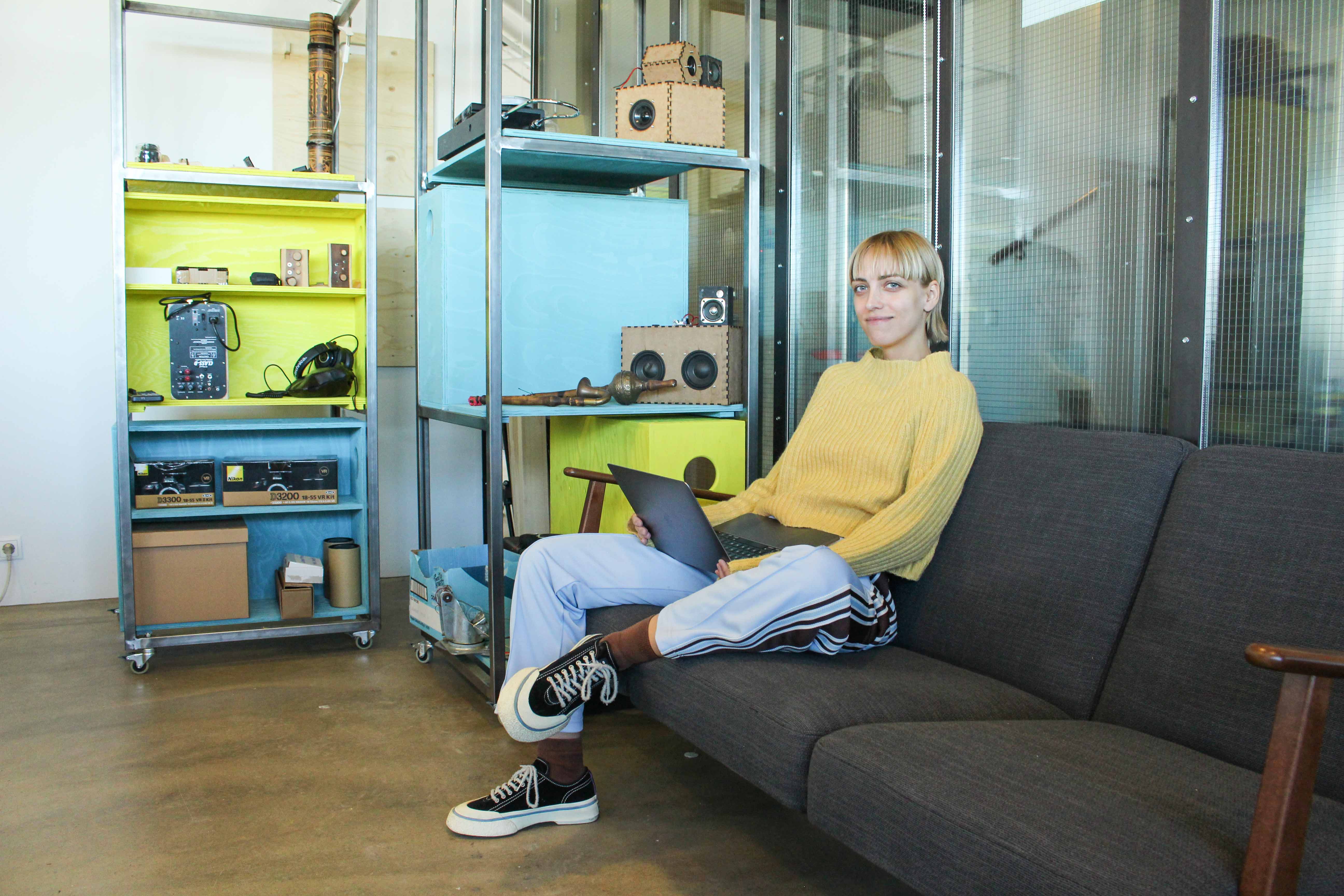 Young woman, dressed in casual yellow and blue clothing, sitting with a laptop in a dark sofa next to yellow and blue shelves.