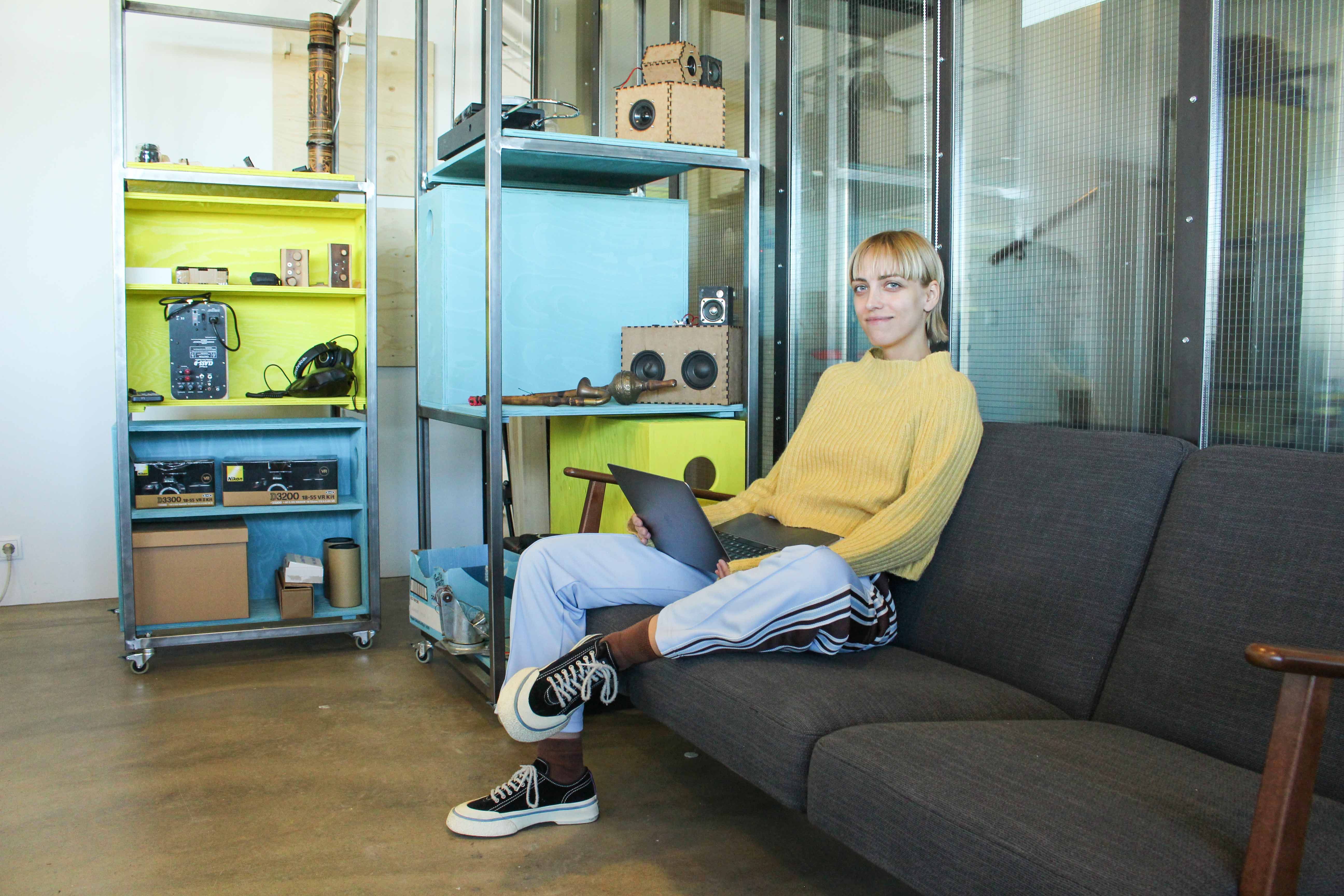 A young woman sitting in a sofa with a laptop, surrounded by yellow and blue shelves