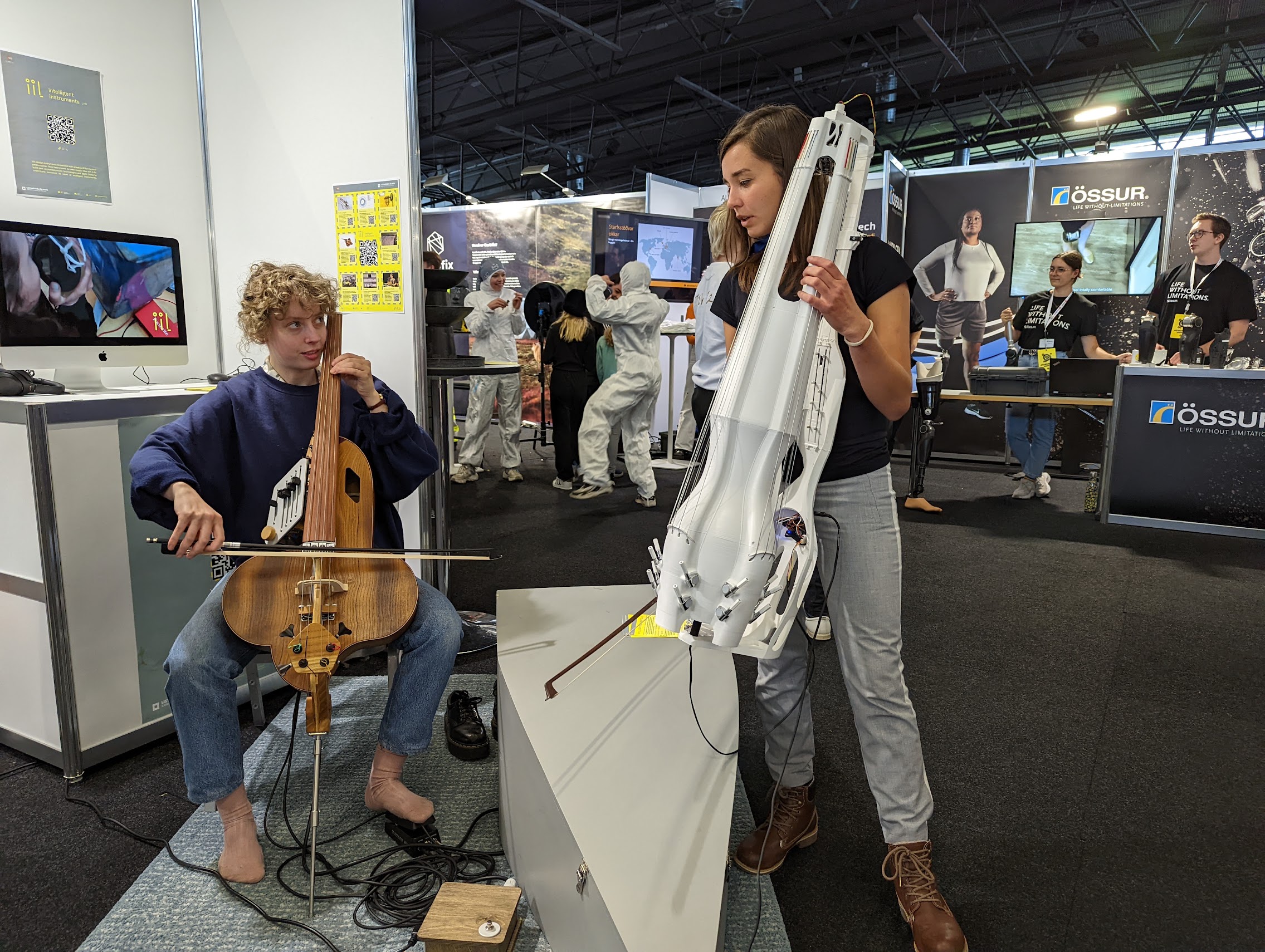 Two young women, each one with an upright instrument in the lap, one playing the halldorophone like a cello, the other one standing with a knurl. In the background, people working at an expo for science.