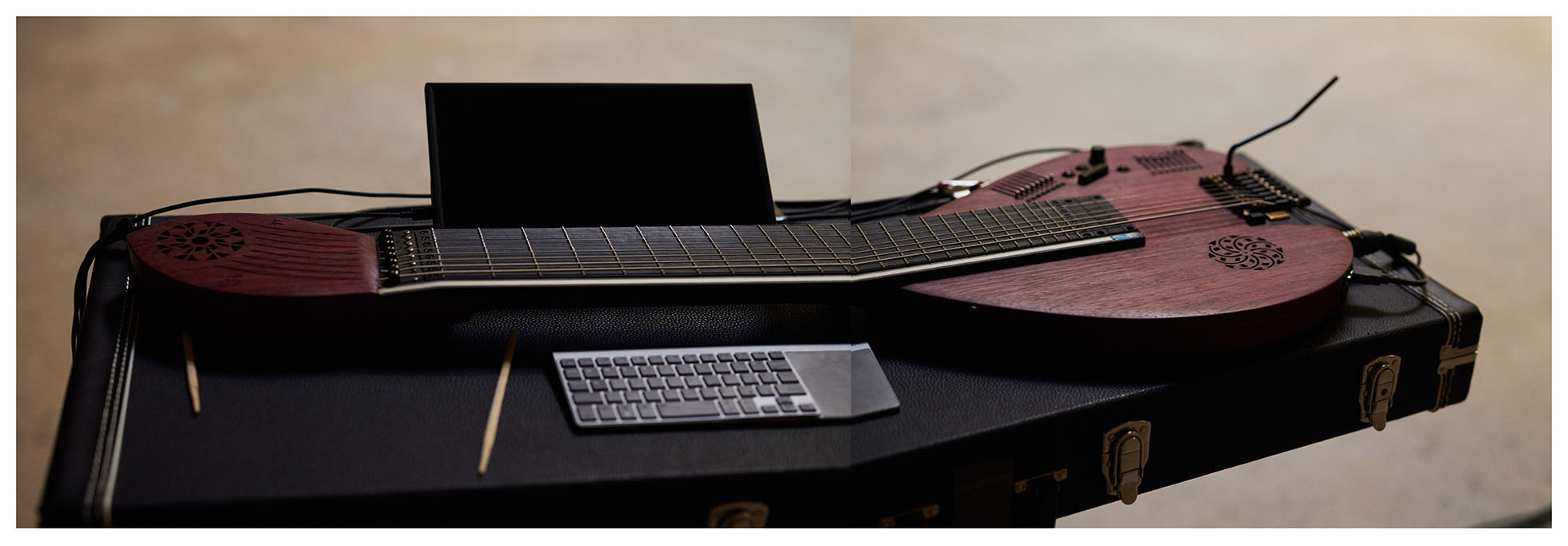 A photo of the Sophtar, an unusual wooden string instrument with embedded electronics. The instrument lies on a black hard case and there are two chopsticks a keyboard and a screen next to it.