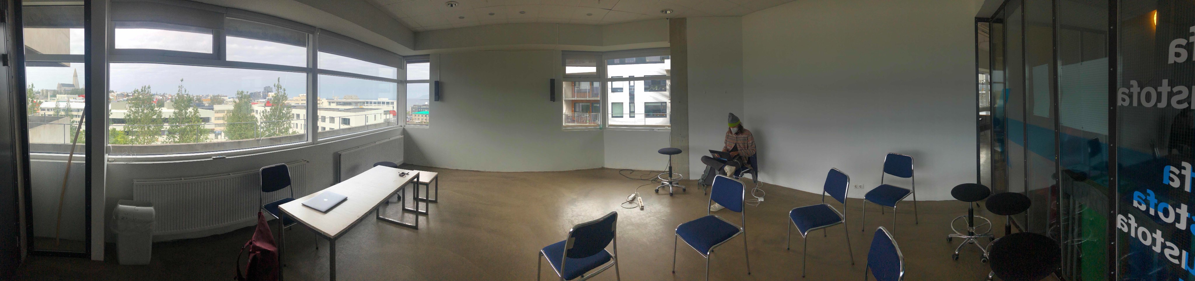 A panorama photo of a large class room what only has one table and a few blue chairs scattered around in a disorganised way. In the middle, a young man sitting in a corner with a laptop.