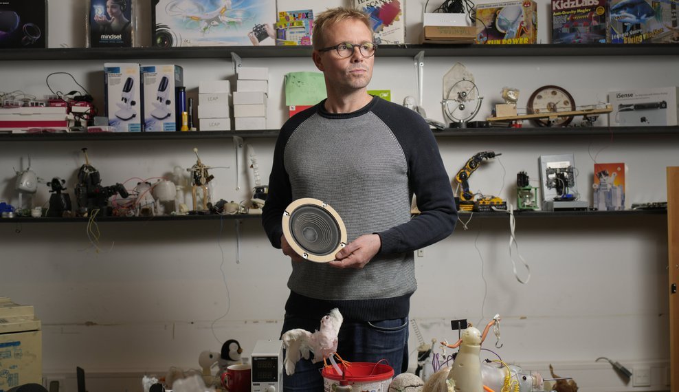 A man standing with a part of a speaker. Shelves in the background.