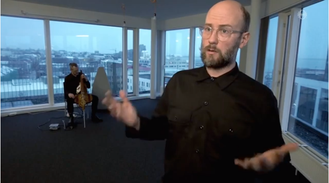 A man speaking to camera. Behind him another man is playing the halldorophone, an intelligent string instrument.