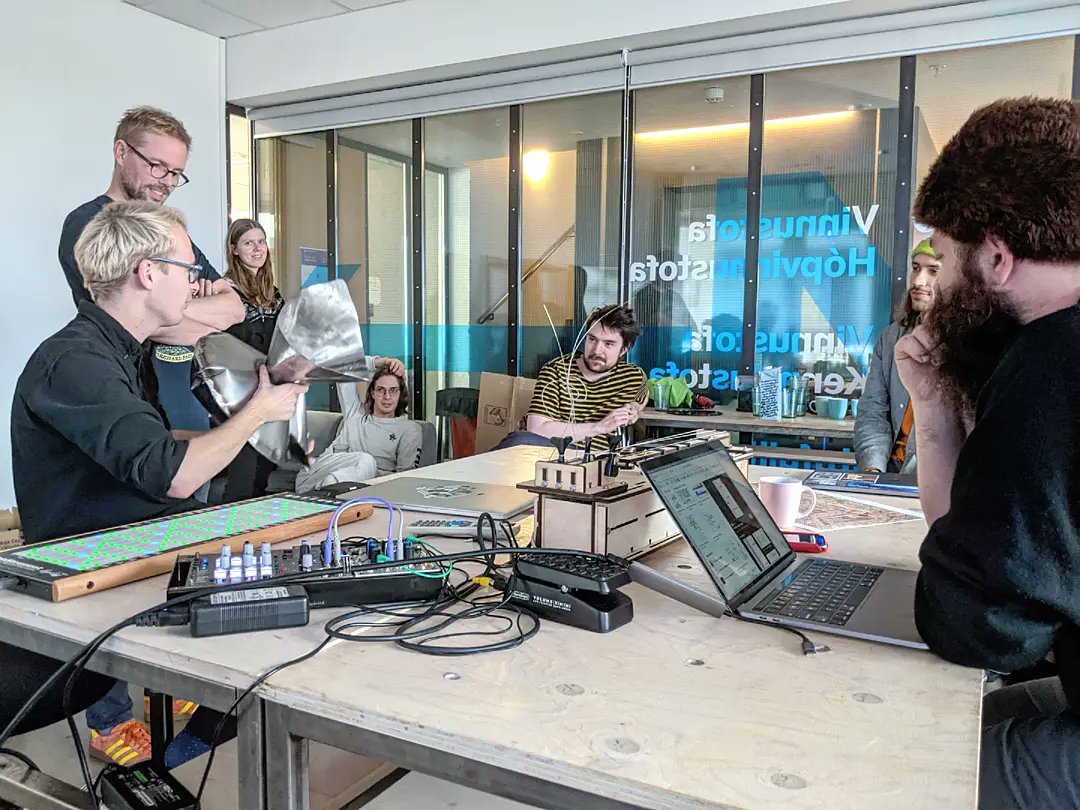 People sitting around a table. Young man holding a metalic prototype of an instrument. Linnstrument, computers, pedal, coffee mug and monochord prototype are among the things on the table.