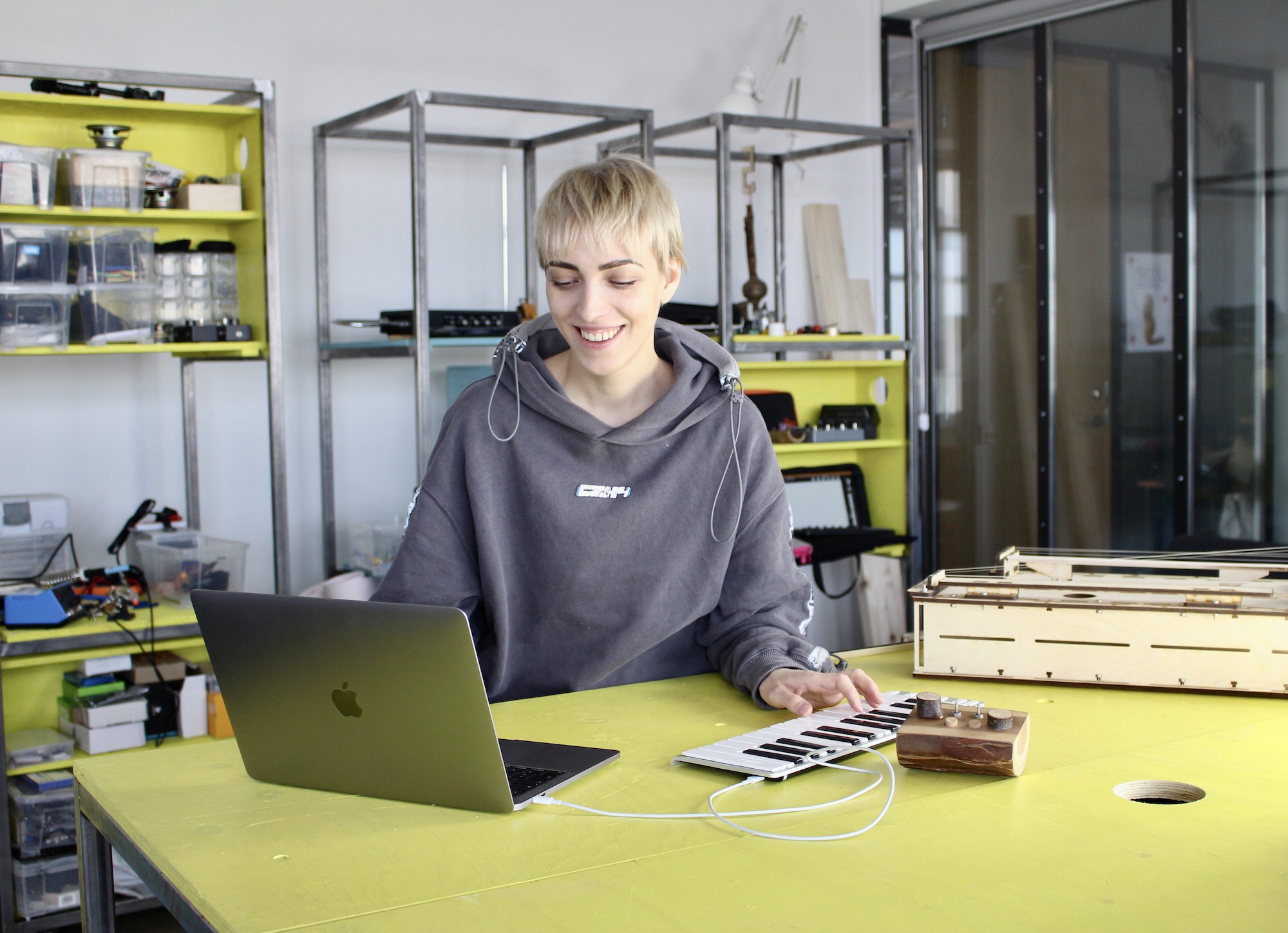 A smiling young woman sitting in front of a laptop and a midi-keyboard. In the background there are yellow and blue shelves with instruments and fabrication devices.